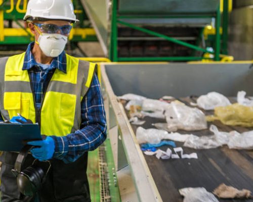 Waste Management Sorting Facility Caucasian Worker Preparing Documentation. Wearing Air Pollution Mask, Eyes Safety Glasses and a Hard Hat.